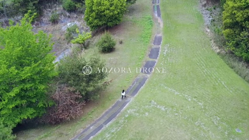 自然豊かな森の中の1本道をゆっくりと歩く女性 ドローン空撮動画素材
